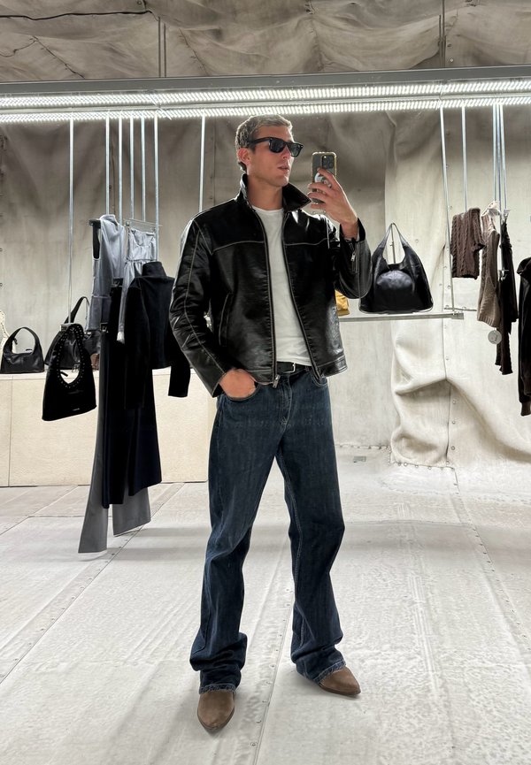 Mann iført svart skinnjakke, hvit skjorte, mørke jeans og solbriller tar en selfie i en butikk med hengende klær og vesker.