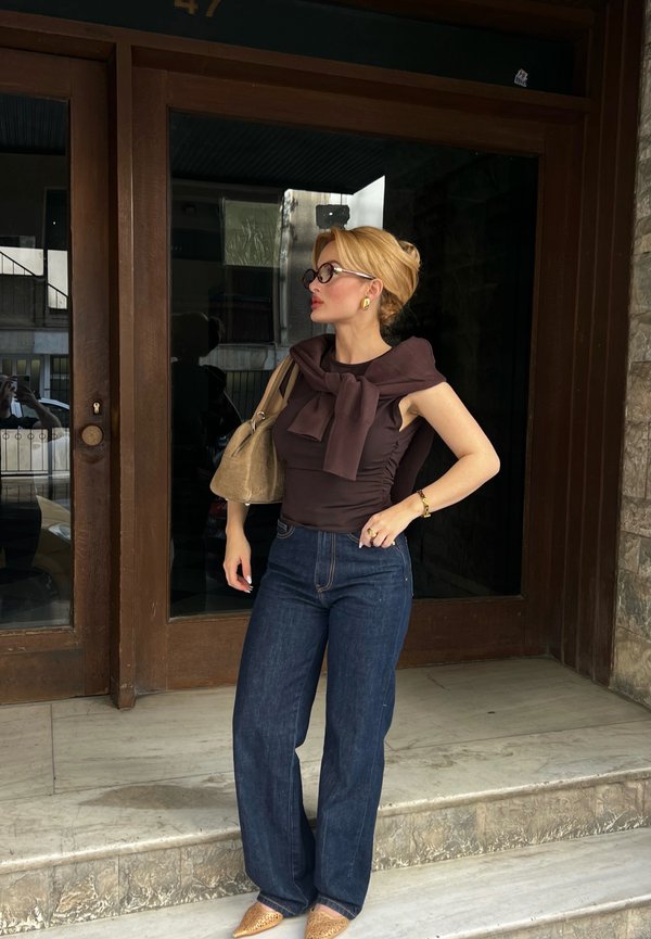 Femme aux cheveux blonds attachés en chignon, portant des lunettes, un haut sans manches foncé avec un pull noué autour des épaules, un jean bleu et des chaussures pointues, debout sur des marches.