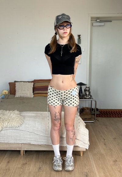 Joven con tatuajes que lleva una gorra, gafas, top corto negro, pantalones cortos de lunares, calcetines y zapatillas, de pie en un dormitorio con una cama y una mesa auxiliar.
