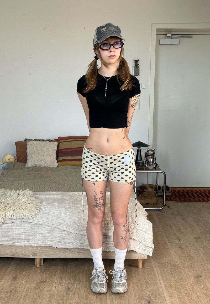 Junge Person mit Tätowierungen, die eine Kappe, Brille, schwarzes Crop-Top, gepunktete Shorts, Socken und Turnschuhe trägt und in einem Schlafzimmer mit Bett und Nachttisch steht.