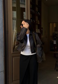 Καφέ δερμάτινο κοντό μπουφάν με αντίθετο ράψιμο, λευκή εφαρμοστή μπλούζα και μαύρο παντελόνι σε φαρδιά γραμμή. Τα αξεσουάρ περιλαμβάνουν γυαλιά ηλίου και τσάντα.