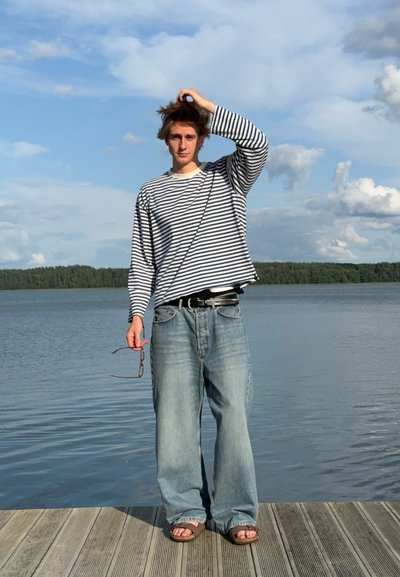 Camisa de rayas de manga larga, vaqueros azules sueltos y sandalias marrones. Sosteniendo unas gafas de sol, de pie en un muelle de madera junto al agua bajo un cielo azul.