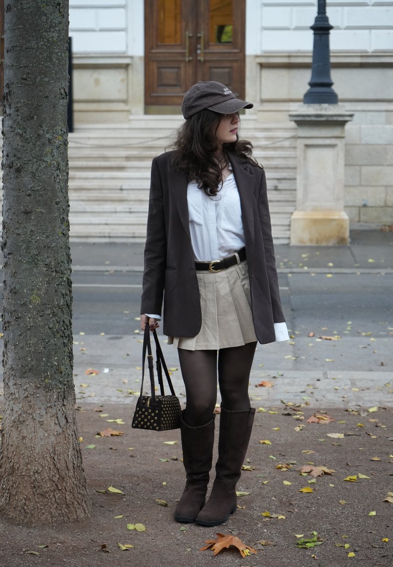 Blazer marron sur chemise blanche, jupe beige, collants noirs, bottes marron montantes jusqu'au genou et sac à main noir à motifs. Debout à l'extérieur près d'un arbre.