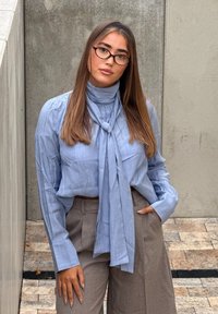 Young woman with long straight hair wearing glasses, a light blue blouse with a necktie, and beige high-waisted pants, standing against a concrete wall.