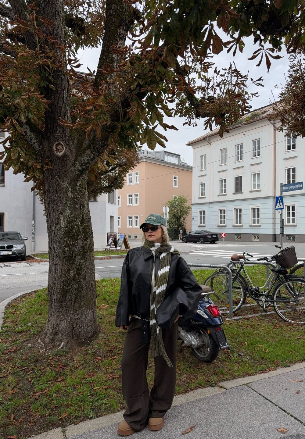 Giacca di pelle nera, sciarpa lavorata a maglia a righe verde e bianco, pantaloni ampi marroni e stivali color cammello, in piedi accanto a un albero con edifici sullo sfondo.