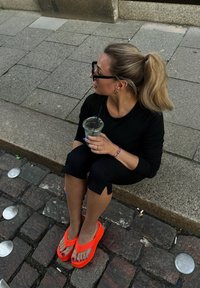 Lyse oransje flip-flops med tykk såle, som ligger på brosteinsbelagt vei, iført svarte cropped bukser og en svart topp; holder en klar drink.
