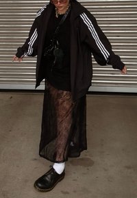 Μαύρο oversized μπουφάν με λευκές ρίγες, φορεμένο πάνω από μια διαφανή μαύρη δαντελ ένια φούστα. Μαύρα παπούτσια, λευκές κάλτσες και μια τσάντα φωτογραφικής μηχανής συμπληρώνουν την εμφάνιση.