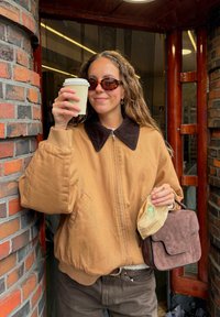 Giacca imbottita marrone con colletto in corduroy, pantaloni marroni scuri, tiene una tazza di caffè e una piccola borsa in suede marrone.