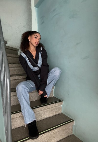 Camisa negra de manga larga bajo un overol de mezclilla azul, combinada con unos pantalones vaqueros de pierna ancha en azul claro y botas negras hasta el tobillo, sentada en unas escaleras.