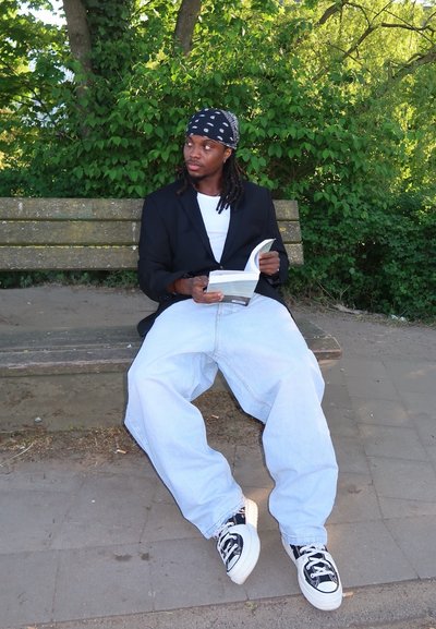 Persona sentada en un banco de madera, con un blazer negro sobre una camisa blanca, vaqueros azules holgados y zapatillas negras, leyendo un libro.