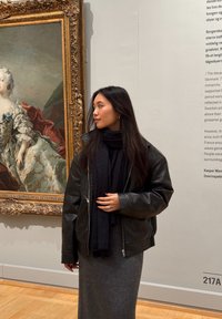 Crna kožna jakna preko tamnog šala, u kombinaciji s sivoj haljini do poda. Prikazuje glatku teksturu i ležeran kroj u muzeju.