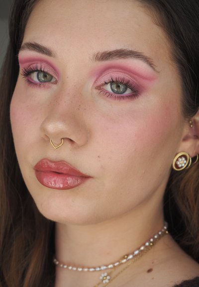 Primer plano de un rostro con sombra de ojos rosa, labios nude brillantes, un aro septal de oro y pendientes con perlas. Pecas visibles en las mejillas.