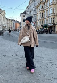 Beige oversized frakke med sorte knapper, sort turtleneck, marineblå hue, stribede bukser og lyserøde platformsneakers, stående på brosten.