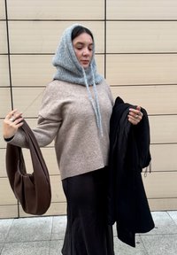 Donna che indossa una sciarpa con cappuccio a maglia grigia e un maglione beige, che tiene un cappotto nero e una borsa marrone, in piedi contro un muro piastrellato.
