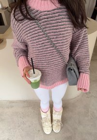 Personne portant un pull rayé rose et gris avec des leggings blancs, des bottes crème, tenant une boisson glacée verte avec une paille, et portant un sac à main gris.