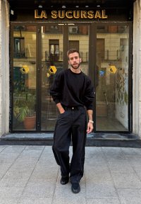 Črna prevelika sweaterka v kombinaciji s širokimi črnimi kavbojkami, ki imajo stranski trak. Okolica vključuje steklena vrata z napisom.
