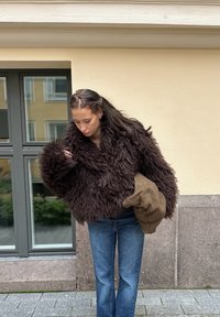 Hnedá bunda z umelého kožúšku s chlpatou textúrou, širokým golierom a nadmerne veľkými rukávmi, kombinovaná s modrými džínsami a hnedou tkanou taškou.