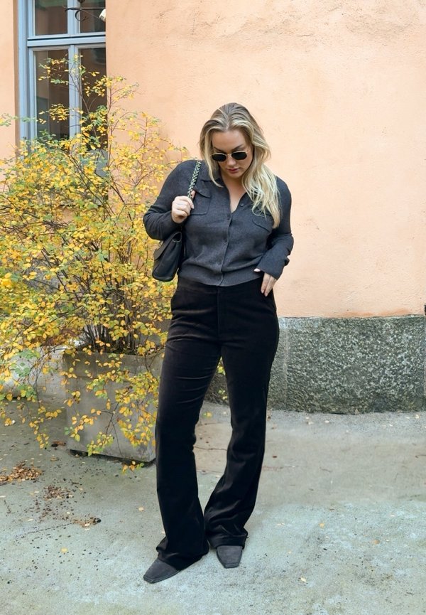 Cárdigan corto de color gris oscuro con dos bolsillos delanteros, combinado con pantalones negros acampanados y zapatos negros de punta afilada, sobre un fondo de pared color melocotón.