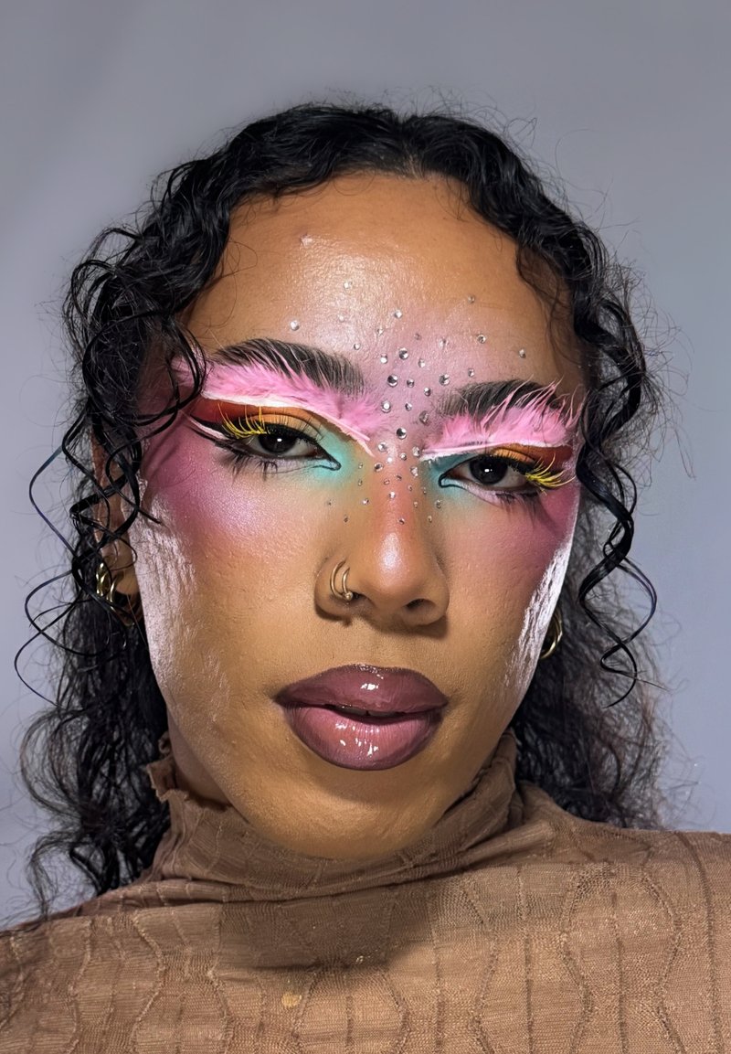 Person with curly hair wears bold makeup with pink feathered eyebrows, multicolored eyeshadow, rhinestones on forehead, glossy lips, and gold hoop earrings.