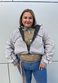 Světle šedá bomberka s černými akcenty, tělový overal s olivovým vzorem, modré džíny, vrstvená šála a výrazné náušnice.