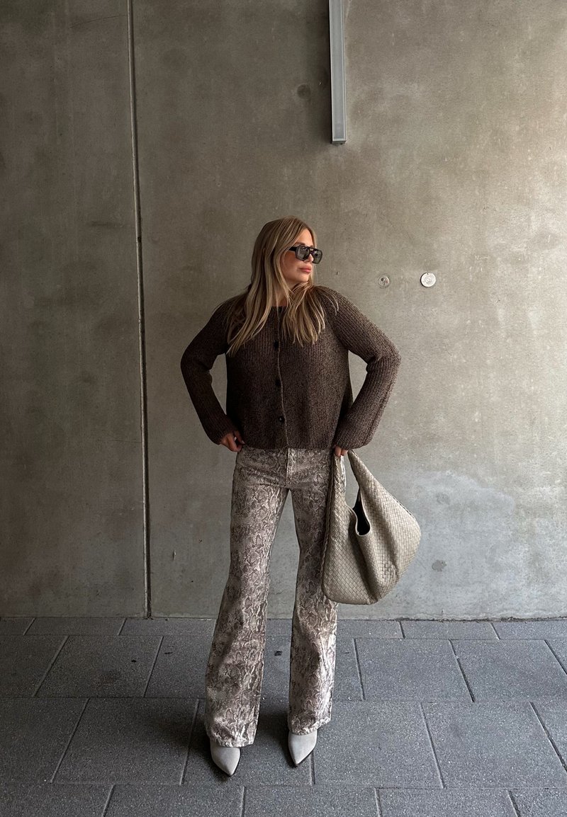 Καφέ πλεκτό cropped cardigan, καρό μπεζ φαρδύ παντελόνι, ανοιχτό γκρι στιλέτο και μια υφή μπεζ τσάντα σε έναν γκρι τοίχο.