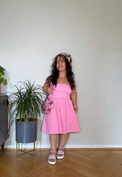 Mujer con el cabello largo y oscuro, vestida con un vestido rosa, una diadema colorida, bolso rosa y sandalias rosas Moschino, de pie en el interior cerca de una planta en maceta sobre un suelo de madera.