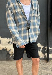 Camicia a quadri blu chiaro e crema con bottoni, due tasche sul petto, indossata sopra una maglietta bianca e abbinata a pantaloni corti neri.