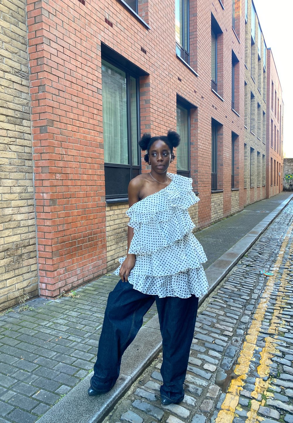 Fehér pöttyös, egyvállas fodros felső laza, sötétkék farmernadrággal párosítva. A háttérben téglából készült épületek és macskaköves utca látható.
