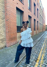 Fehér pöttyös, egyvállas fodros felső laza, sötétkék farmernadrággal párosítva. A háttérben téglából készült épületek és macskaköves utca látható.