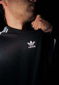 Černá atletická mikina s bílým logem Adidas, strukturovanou tkaninou, kulatým výstřihem a třemi bílými pruhy na rukávech.