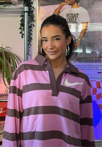 Jeune femme portant des boucles d'oreilles anneaux et un anneau au nez, habillée d'un t-shirt Nike rayé rose et marron, debout à l'intérieur avec des plantes et une affiche derrière elle.