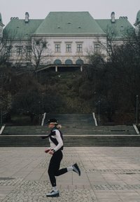 Atleet in zwarte legging en wit topje, rennend op een stenen trottoir met kasseien, op de achtergrond een groot gebouw met een groen dak.