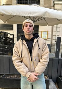 Beige hoodie met zwarte voering, rits aan de voorkant en logo patch. Draagt over een zwarte sweatshirt, in combinatie met lichtblauwe jeans en een beige beanie.
