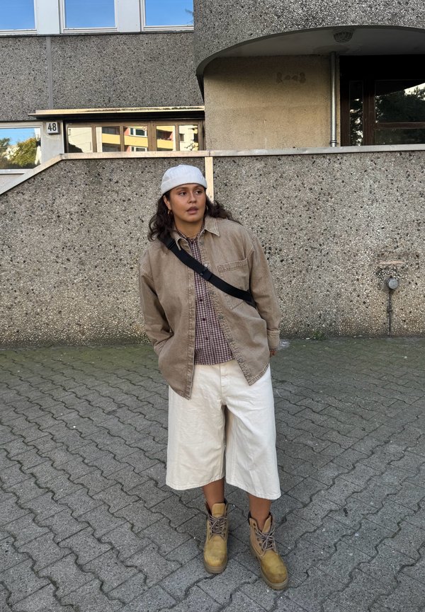 Persona joven vistiendo chaqueta beige, sombrero blanco, camisa a cuadros, pantalones cortos blancos holgados y botas marrones, de pie en una zona pavimentada frente a un edificio de hormigón.