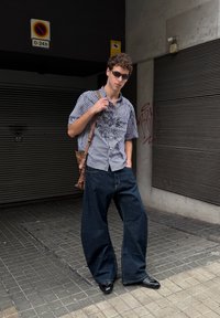 Rutete skjorte med kort erme, baggy mørke denimjeans og svarte spisse sko. Brun skulderveske og solbriller fullfører antrekket.