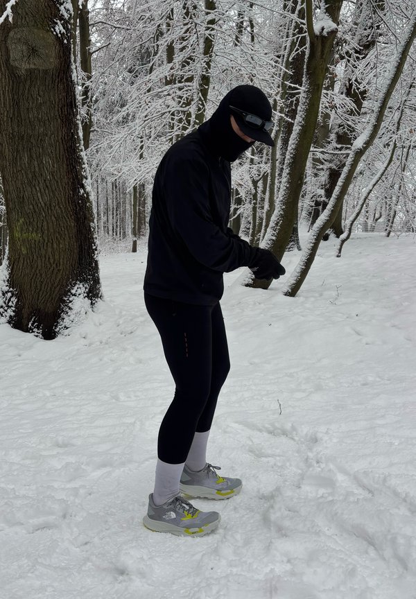 Person in schwarzer Winterkleidung, Sonnenbrille und Mütze steht in einem mit Schnee bedeckten Wald mit kahlen Bäumen im Winter.