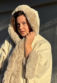 Roomkleurige mantel met capuchon en een zachte nepbontafwerking. De stof lijkt fluweelachtig, met een losse pasvorm en een grote capuchon.