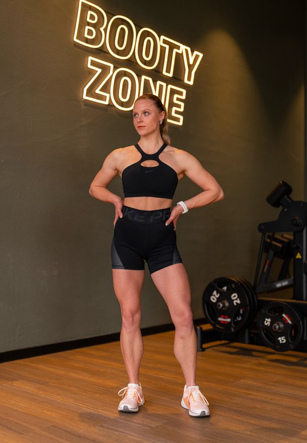 Fit vrouw in zwarte sportkleding staat met handen in de zij onder het neonbord "BOOTY ZONE" in een fitnessruimte met gewichten op de achtergrond.