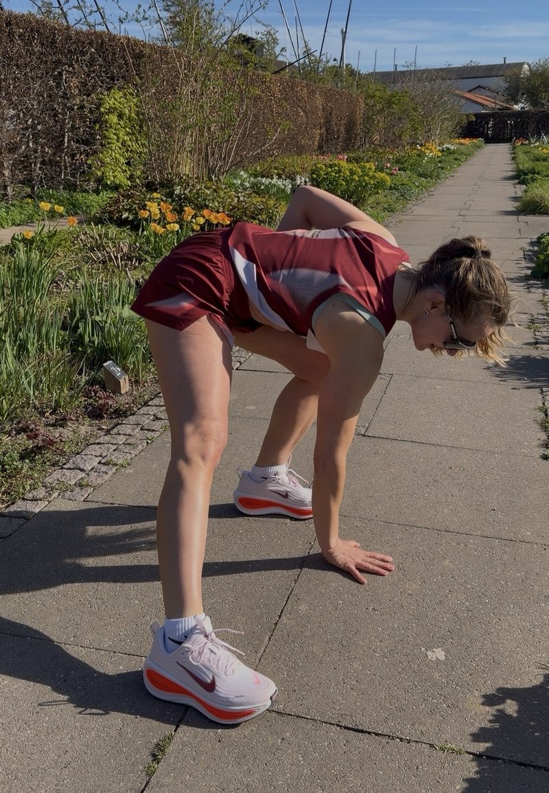 Femme en tenue de sport se penchant en avant avec une main sur le pavé d'un chemin de jardin bordé de fleurs et de verdure.
