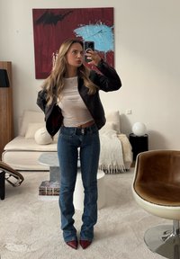 Žena s dugom plavom kosom koja nosi crnu kožnu jaknu, bijelu majicu, plave traperice i crvene cipele sa špicom, snima selfie u ogledalu u dnevnoj sobi.