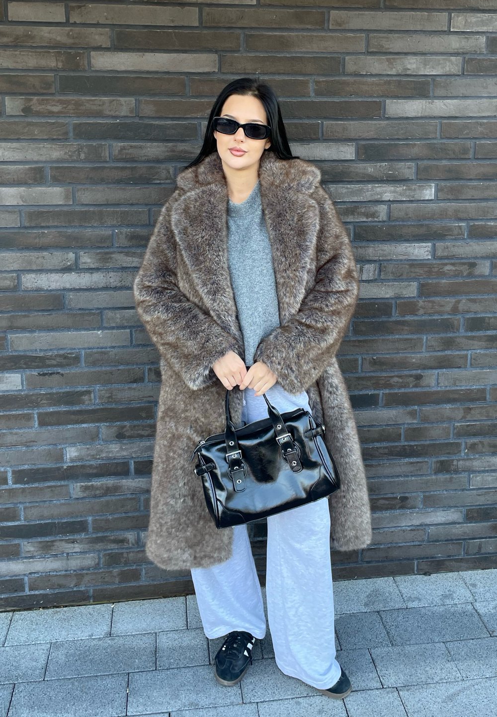 Καφέ ψεύτικη γούνα παλτό με φαρδιές πέτες, γκρι πουλόβερ, ανοιχτό γκρι παντελόνι και μια μαύρη γυαλιστερή τσάντα χειρός με μεταλλικές λεπτομέρειες, στέκεται μπροστά σε έναν σκοτεινό τοίχο από τούβλα.