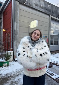 Jeune femme aux yeux fermés portant un manteau en fourrure blanc à taches noires et un pull en tricot, debout dehors dans une rue enneigée près de bâtiments industriels.