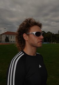 Óculos de sol desportivos com armações prateadas e lentes escuras, usados por uma pessoa com uma camisa de manga longa preta com riscas brancas nos ombros.