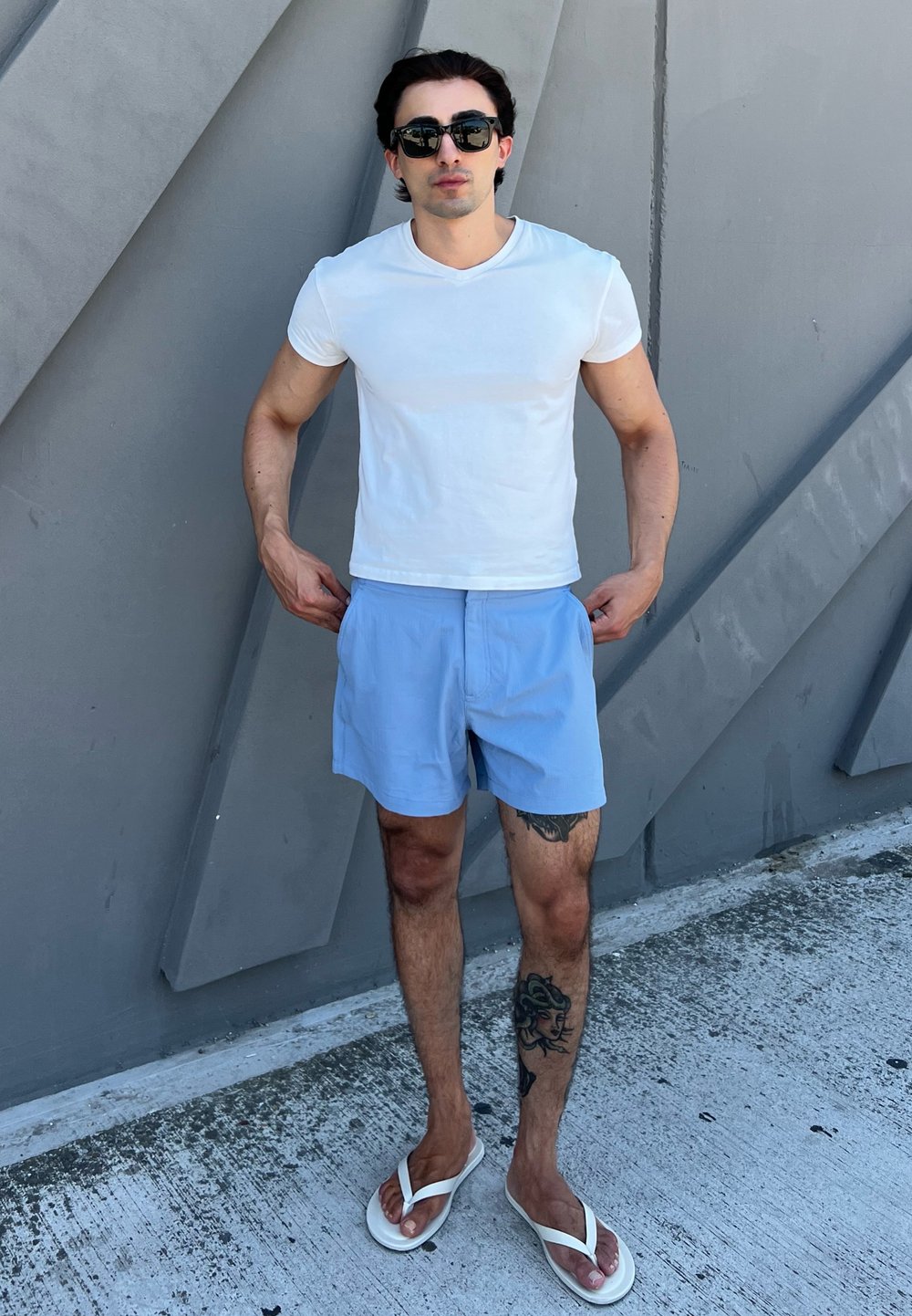 Camiseta branca justa, calções azul claros e chinelos brancos. O fundo apresenta padrões geométricos cinzas. Tatuagem visível na perna.