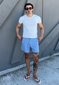 Camiseta branca justa, calções azul claros e chinelos brancos. O fundo apresenta padrões geométricos cinzas. Tatuagem visível na perna.