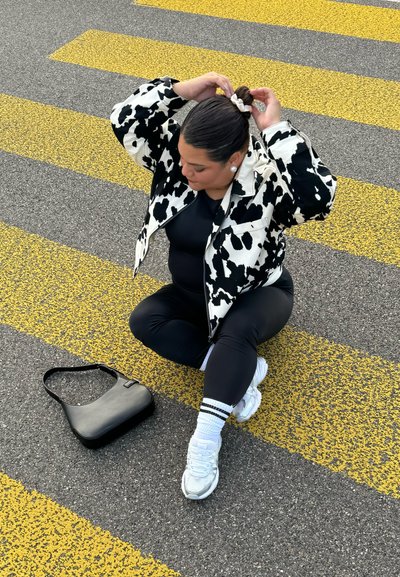 Chaqueta de cremallera con estampado de manchas de vaca en blanco y negro sobre un conjunto negro, sentada en un pavimento gris con rayas de paso de cebra amarillas, sosteniendo un coletero.