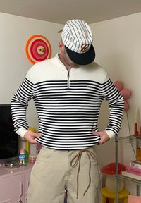 Maglione a righe bianche e nere con cerniera e collo alto. Pantaloni beige con coulisse. Indossa un berretto a righe.