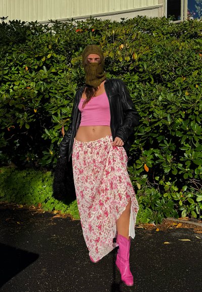 Balaclava de punto verde, chaqueta de cuero negra, top corto rosa, falda larga de flores y botas altas rosa, posando contra un fondo verde.