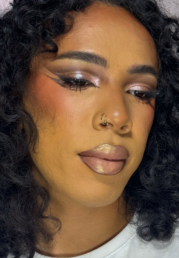 Personne aux cheveux noirs bouclés et anneaux de nez dorés porte un maquillage des yeux dramatique avec paillettes, eye-liner ailé et rouge à lèvres métallique doré.