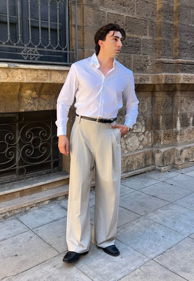 Hombre de pie en la acera junto a un muro de piedra, vistiendo camisa blanca de vestir, pantalones beige, zapatos negros y reloj de pulsera, mirando de lado.